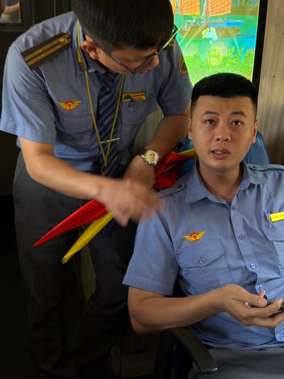 Two men in blue, button-down shirts, one with epaulettes, are indoors. Both wear trousers. One wears a wristwatch and has his hand extended, holding a red and yellow object. The other man looks directly at the camera. Behind them is a screen with what appears to be the cityscape of Huế, Vietnam, showing boats and buildings along the river.
