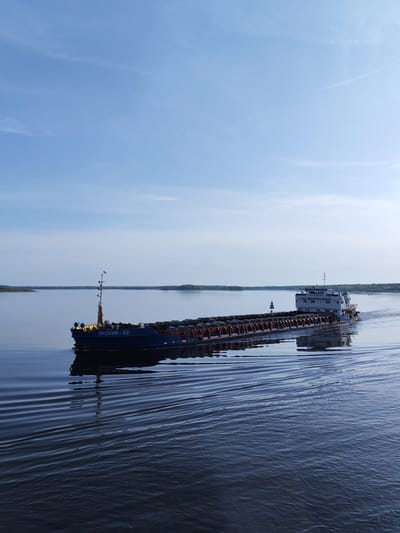 The image shows a cargo ship, named "ОКСКИЙ - 62", moving on a calm body of water. The ship appears to be carrying cylindrical cargo. The sky is clear with a few wisps of clouds. The shoreline is visible in the background with green vegetation. The location is likely near Shelomovo, Russia.