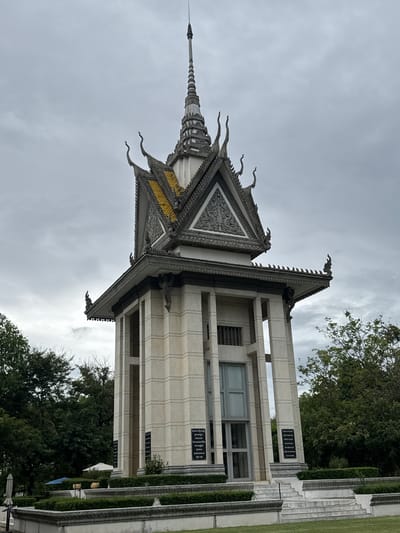The image shows the Choeung Ek Genocidal Center Memorial Stupa in Khan Dangkao, Cambodia. The stupa is a tall, multi-tiered structure with a distinctive spire. Its exterior is made of light-colored stone with decorative carvings and ornate roof details. The stupa has an open, columnar design on its lower levels, revealing a central glass-enclosed structure. Black plaques are visible on the exterior walls. The stupa is surrounded by manicured greenery and steps leading up to its base. In the background, trees and a cloudy sky are visible. The overall impression is solemn and reflective.
