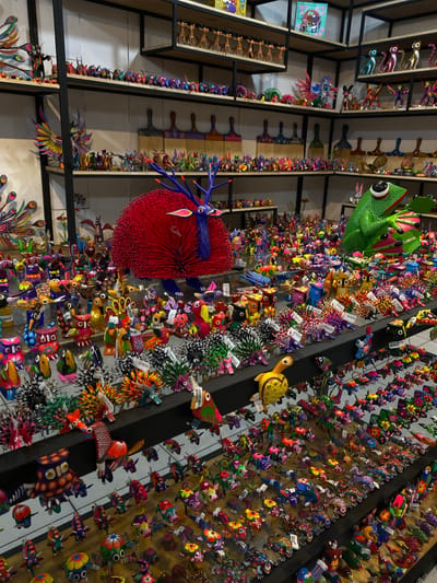 The image depicts a vibrant display of colorful alebrijes (Mexican folk art sculptures of fantastical creatures) in Oaxaca City, Mexico. The alebrijes, primarily animals and mythical beings, are densely packed on shelves and wall-mounted displays. They are varied in size, from small figurines to larger, more elaborate pieces. The shelves are constructed of dark metal frames and wood panels, creating a grid-like structure for displaying the alebrijes. Price tags are visible on many of the items, indicating they are for sale. The scene appears to be a shop interior, showcasing the artistic craftsmanship and cultural heritage of the region.