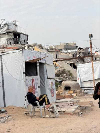 The image captures a scene in Jabalia, Palestinian Territory, characterized by extensive urban destruction and the presence of temporary shelters. In the midground, a middle-aged to elderly man with short grey hair and a beard sits on a light-colored plastic chair, reading an open book or document. He wears a dark long-sleeved top and dark track pants with yellow stripes; his bare feet rest on the inverted seat of a second light-colored plastic chair placed in front of him. To his left, a temporary structure made from white tarpaulin panels features an open dark entryway and visible Arabic script in red and black ink. Another similar white tarp structure is partially visible to the right. Between these structures, a small, dome-shaped concrete or mud-brick structure resembling an outdoor oven stands on a stone base. A tall, rusty metal pole with a dark attachment at its top is positioned on the far right, where a partial view of another person's dark-clothed arm and hand is visible. The foreground consists of uneven light brown dirt and sand with scattered debris, leading to a slightly elevated paved area. The background reveals widespread devastation: multiple multi-story buildings are reduced to rubble and twisted metal, with some structures partially collapsed. The sky is overcast and uniformly light grey.