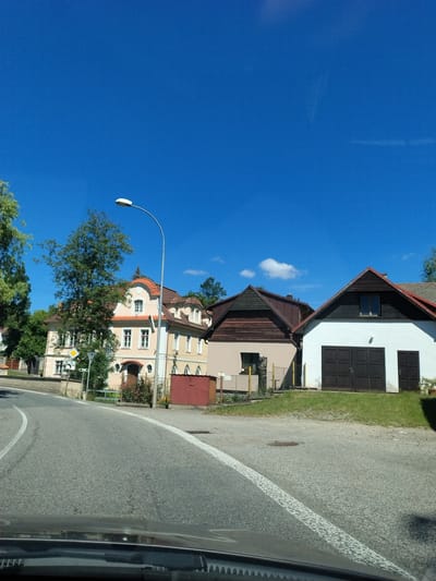 The image shows a street scene in Čkyně, Czechia. On the right side of the road, there are two buildings: one is a white garage with a large brown door and a smaller brown door on the right, and the other is a beige building with a brown roof. In the background, there is a light yellow building with an orange-tiled roof, featuring multiple windows and an arched doorway. A street lamp is visible on the left side of the road, with a clear blue sky above and some green trees and foliage. The road itself is paved and has white lane markings. The foreground shows the dashboard and windshield of a vehicle, indicating the photo was taken from inside a car.