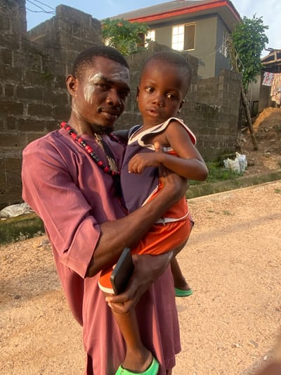 In Afikpo, Nigeria, a man carries a child. The man wears a maroon robe, a necklace of black and red beads, and has white paint on his forehead. He holds a black phone in his hand. The child wears an orange and blue tank top and green shoes. They are standing outside near a stone building on a sandy pathway.