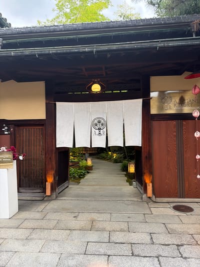 The image presents the main entrance to Akagane Resort, located in Kyoto, Higashiyama, Japan. The entrance is framed by a traditional Japanese wooden structure featuring dark brown pillars and beams, surmounted by a grey tiled roof with exposed gutters. A prominent white fabric "noren" curtain, split into four panels, hangs centrally in the open doorway, displaying a black circular logo with an "A&R" monogram and a stylized peacock feather motif. Above the noren, a single yellow spherical lantern is suspended within an octagonal wooden frame.

To the right of the entrance, a horizontal gold-toned sign is mounted above a dark wooden panel, clearly stating "AKAGANE RESORT KYOTO HIGASHIYAMA 1928" alongside the "A&R" logo. A string of pink, multi-lobed decorative ornaments hangs vertically from the right side of the entrance structure. On the left side, a partially visible white sign with black text reads "WELCOME TO OUR WEDDING," positioned next to a small arrangement of dark red flowers.

The forecourt leading to the entrance is paved with irregular grey stone blocks. A stone pathway extends beyond the threshold into a meticulously maintained garden, visible through the open entrance. This garden is lush with various green foliage, including tall bamboo, and is illuminated by two square, patterned ground lanterns positioned along the pathway. The background reveals an overcast sky and the upper parts of green trees. No people are visible in the scene. A circular metal manhole cover is present on the paved ground to the lower right.