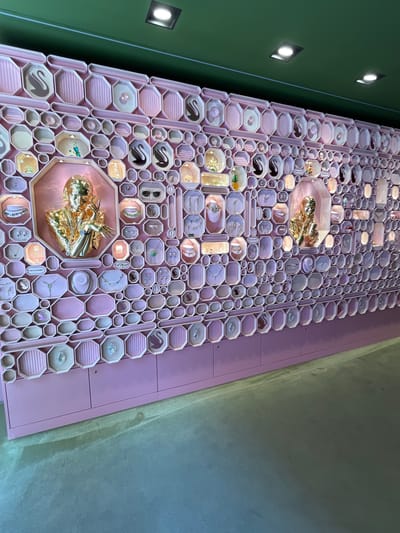 The image depicts a wall display of Swarovski jewelry inside a store. The display is a matrix of pink, octagonal and rectangular compartments, each containing a piece of jewelry, ranging from earrings and necklaces to small sculptural objects. Two larger, prominently placed compartments hold gold-colored sculptures. The display is lit by overhead lights, creating highlights and shadows on the jewelry. The store is located in Verona, Italy. The wall to the left of the display is painted green, while the wall to the right is a textured gray.