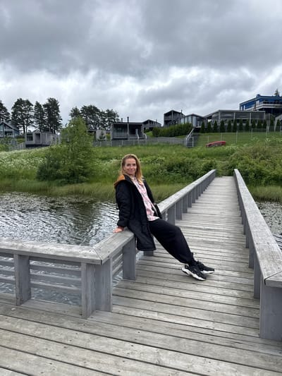 A woman is sitting on the edge of a wooden pier that extends over a body of water. She is wearing a black quilted jacket with a tan hood, a pink and white checkered shirt, and black pants. She has on black athletic shoes with white soles. The pier is made of weathered gray wood planks and has a railing on both sides. The water is dark and rippled. In the background, there is lush green grass, trees, and several modern houses with dark exteriors and large windows. The sky is overcast and gray. The location is described as the city of Mars, Russia.