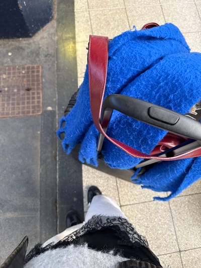 A high-angle view depicts a partial individual and luggage within an urban or transit environment in Newcastle upon Tyne, United Kingdom. The observer's feet, clad in black shoes and a light-colored garment with black lace trim, are visible at the bottom. To the right, a wheeled luggage item is covered by a vibrant royal blue, thick, textured textile, consistent with a blanket or scarf. A deep red leather strap, likely from a handbag or shoulder bag, is draped over the luggage's black extendable handle and across the blue textile. The ground features light-colored square paving tiles, transitioning to a dark grey or black curb on the left. A square metal drainage grate is embedded in the darker ground section on the far left. Natural light illuminates the scene.