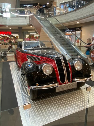 This is a vertical, medium-shot photograph taken indoors, likely in a shopping mall or exhibition space in Łódź, Poland, as indicated by the text "LODZKA" and the mall-like architecture. The primary subject is a vintage two-tone black and red car, possibly a convertible due to its open top. The car is prominently displayed on a metallic checkered platform. A sign attached to the car reads "AUTO-MOTO-MUZEUM" and "Oltimet Kaptive," suggesting it is part of a museum exhibit.
In the background, an escalator is visible, with several people casually walking or standing on it. One woman with blonde hair is looking towards the car, while another person is partially visible behind her. Below the escalator, further into the mall, shops are visible, including one with a sign that says "BETTY BARCLAY." Another sign displays "NORTH" in large red letters, possibly indicating a department store or the name of the mall. A red sign with white text also shows "30%," indicating a sale. The overall atmosphere appears to be casual and commercial, with no strong indicators of time of day or weather. The lighting is artificial, typical of an indoor shopping environment.