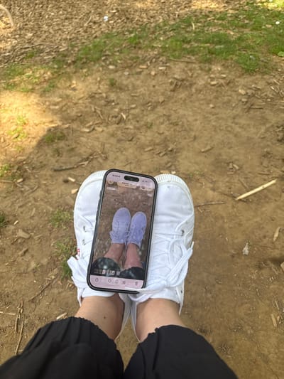 The image, taken in Pétange, Luxembourg, presents an overhead view of a person's legs and feet positioned on an outdoor dirt path. The person is wearing black pants and clean white canvas lace-up shoes, one of which has a small red "LEVI'S" tag visible on its side.
A silver and black smartphone is held directly above the feet, its screen facing upwards. The phone's display shows a photograph of a person's feet, wearing light purple or lavender canvas shoes, also appearing to be on a dirt ground. This creates a recursive "photo within a photo" effect.
Notable details on the phone screen include a timestamp of "16:03" (4:03 PM) and the word "Aujourd'hui" (Today), confirming the time of day. A "LIVE" button is visible, along with a "5G" network indicator and various camera mode icons.
The setting is an unpaved natural environment, likely a path or trail, characterized by light brown soil, patches of green grass, and scattered organic debris such as small twigs and dry leaves. The bright, natural illumination suggests it is daytime. The scene captures a casual moment of photographing footwear outdoors.