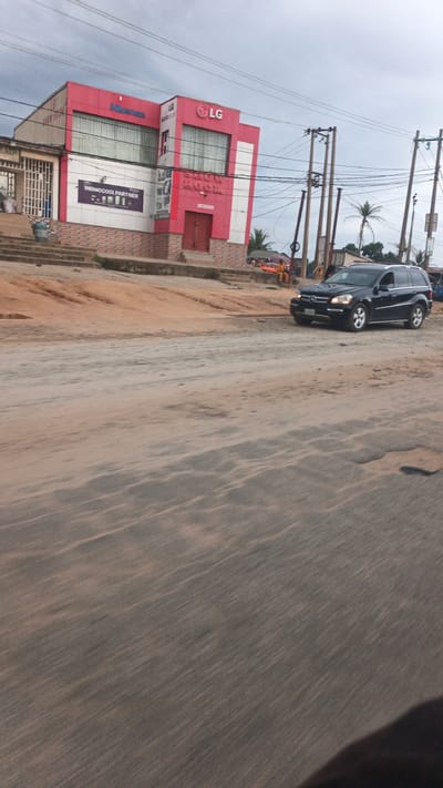 The image depicts a street scene in Suleja, Nigeria. A red and white building with prominent "LG" and "Hisense" signage dominates the left side of the frame, functioning as a showroom. A dark-colored SUV is parked on the right side of the road, facing left. The road surface is uneven and appears to be unpaved in some areas, with visible sand and dust. Electrical poles and wires are present in the background. A few individuals are visible in the distance, near the building. The sky is overcast.