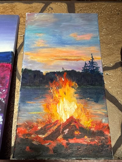 The image displays a vertical canvas painting of a campfire scene. The painting is set against a backdrop of a twilight sky with streaks of orange, pink, and blue, suggesting a sunset or sunrise. Below the sky is a dark silhouette of a forest, with a tall, pointed evergreen tree standing out against the treeline. A body of water, likely a lake or river, reflects the sky and the dark forest. In the foreground, a vibrant campfire blazes with thick impasto strokes of yellow, orange, and red, illuminating the logs and embers. The base of the painting is dark, suggesting the ground around the campfire. The painting is positioned on a textured surface, possibly a table with a stone or tile pattern, and another canvas painting with purple and pink hues is partially visible on the left. There is no indication of a specific location in Naro-Fominsk, Russia.