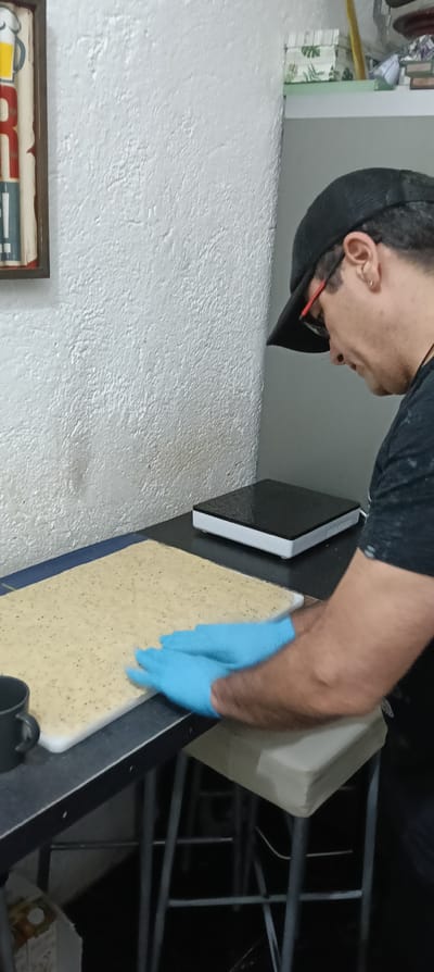 An individual is depicted in an indoor setting, likely a kitchen or food preparation area, in Ripollet, Spain. The subject, a male, is viewed in profile from the right, wearing a black baseball cap, red-framed eyeglasses, a black t-shirt, and blue disposable gloves. He possesses a small metallic earring in his visible ear. Both gloved hands are pressed down onto a rectangular, light-colored, thin substance containing small dark inclusions, resembling dough or a similar food item. This substance rests on a white cutting board, which in turn sits on a dark countertop or table. To the immediate left of the cutting board, a black mug is present. Further right on the countertop, a white electronic device with a black top surface is visible. The background features a textured white wall. A brown-framed sign, displaying partial red and yellow lettering (appearing to be "ER"), is mounted on the wall to the left. A dark cabinet or shelving unit stands to the right, and a shelf above it holds various objects, including a white box with a green leaf pattern. Below the countertop, the metal frame and a padded light-colored seat of a stool are discernible. The floor is dark.