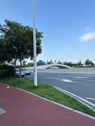 In Haizhu District, China, a clear blue sky with scattered clouds overlooks a modern urban scene. A paved road with white directional arrows dominates the foreground, bordered by a red jogging path and a strip of green grass. Two silver sedans are parked on the grass to the left, partially obscured by a streetlamp. In the background, a white, arching structure spans across the road, adorned with golden Chinese characters. Beyond the structure, trees and modern buildings rise against the sky. A blue traffic sign with white lettering is visible on a pole. The overall impression is one of a well-maintained urban environment on a bright, clear day.