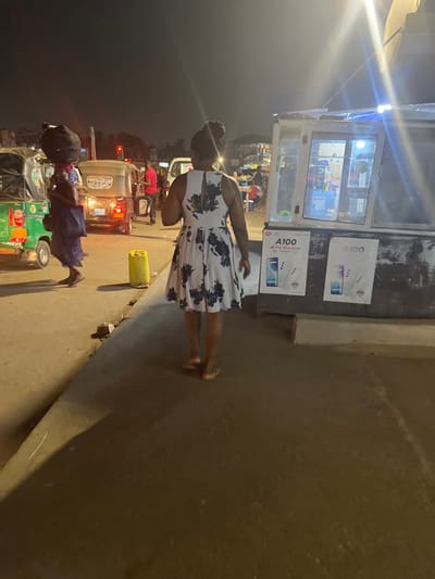 A woman in a floral dress stands on a sidewalk next to a small shop. The shop has product advertisements and bright lights. A busy street with traffic is visible in the background, including cars and small rickshaws. Another person walks along the road with what appears to be a vendor's cart. The scene is illuminated with streetlights, suggesting it is nighttime.