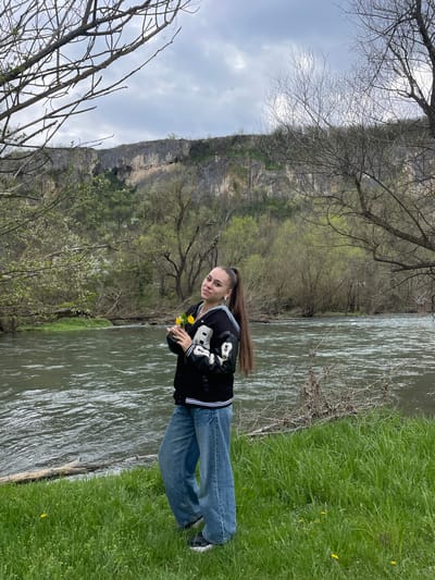 The image shows a woman standing next to a river in what appears to be a natural setting. A cliff face can be seen in the background. The woman is wearing a black jacket and jeans. She is holding yellow flowers. The environment includes a river with visible water movement, grassy banks on one side, and trees. The location is identified as Barkach, Bulgaria.
