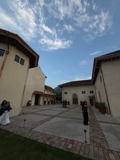 The image shows an outdoor courtyard area with several people present, located in San Pedro Garza García, Mexico. The architecture of the buildings surrounding the courtyard is European-inspired, with beige walls, copper rain gutters, and red tile roofs. In the lower part of the image, two women are standing together, dressed in dark shirts and light-colored skirts. Near the center of the image, further up the courtyard, another woman is standing, holding her phone up as if taking a picture. There are brick pathways that intersect with concrete walkways. At the end of the courtyard, closer to the buildings, a group of people is standing near a statue. The sky is blue with wispy clouds.