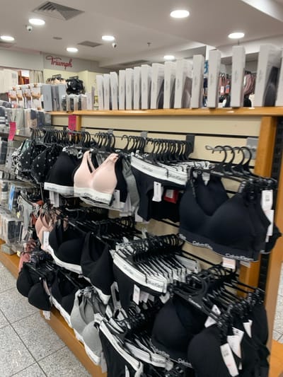 The image displays a retail store setting, specifically a lingerie section, as seen from a slightly elevated perspective. The main subjects are numerous bras and underwear sets, predominantly from the Calvin Klein brand, neatly arranged on black plastic hangers across multiple racks. The garments are mostly in black, grey, white, and light pink.
The activity taking place is the display of merchandise for sale within a store. The items are presented in an organized manner, ready for customers to browse.
Notable details include the prominent "Calvin Klein" branding visible on the elastic bands of the underwear and bras. On the left side of the image, stacks of white product boxes are visible on shelves. Further in the background, a red sign with the word "Triumph" indicates another brand or section within the store. The store is well-lit with recessed artificial lighting from the ceiling, suggesting daytime hours. The floor is tiled. No people are visible in the image. The scene conveys an orderly and commercial atmosphere typical of a department store or a specialized apparel shop in Marousi, Greece.