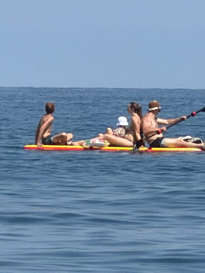The image depicts four people on a paddleboard in the sea. The perspective is from above, looking down at the paddleboard which is oriented vertically in the frame. The water is a deep blue and appears calm with slight ripples.
The people are arranged along the length of the paddleboard. The person at the top of the board, likely the driver, is a man with a helmet and is holding a paddle. Below him, a woman is also sitting on the board. Further down, another person, possibly a child due to their size, is wearing a white hat. The person at the bottom of the paddleboard is a man lying down, holding onto the board. All individuals appear to be wearing swimsuits. The paddleboard itself is brightly colored with red and yellow accents.
The scene suggests a recreational activity, likely a summer day given the attire of the individuals and the clear blue sky, although the sky is mostly out of frame. The lighting suggests it is daytime, possibly late morning or afternoon. There are no visible signs or text in the image. The setting is a body of water, consistent with a coastal area. The people's postures suggest they are either enjoying a leisurely paddle or perhaps are in the process of maneuvering the board.