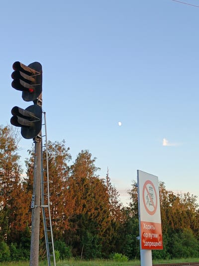 This image depicts a railway crossing in Losava, Belarus, under a clear sky with the moon visible. The scene is set during the late afternoon or early evening, with the sun casting a warm glow on the trees in the background.
The primary subjects are a railway traffic signal and a warning sign. The traffic signal, positioned on a metal pole with a ladder, shows a red light illuminated, indicating that trains are present or approaching and passage is restricted. The warning sign, featuring a red circle with a diagonal line through it, displays an image of a person walking on train tracks, signifying that walking on the tracks is prohibited. Below this symbol, text in Russian reads "Ходить по путям запрещено!", which translates to "Walking on the tracks is prohibited!".
Other details include a marker on the pole labeled "H 3", likely indicating the location or a specific point along the railway. The background is filled with dense green trees, some of which are illuminated by the setting sun, giving them an autumnal hue. The overall impression is one of caution and safety regulations at a railway crossing, emphasizing the danger of walking on the tracks. The presence of the moon suggests a transition from day to night, contributing to a serene yet cautionary atmosphere.