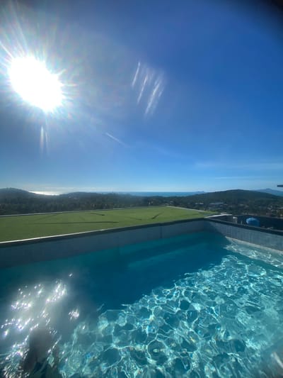 This media file captures an elevated view of an infinity pool with sparkling, clear blue water. The pool is bordered by a gray tiled edge and a strip of artificial turf. Beyond the pool, a lush, green hillside covered in trees and dotted with buildings slopes down towards the ocean. In the distance, a hazy coastline and the sea are visible under a bright, clear blue sky.
The sun is prominently featured in the lower left quadrant of the image, casting a strong lens flare and reflecting off the water's surface, creating a shimmering effect. There are faint, white streaks in the sky that could be contrails from an aircraft.
The image appears to be taken during the daytime, likely late morning or early afternoon, given the intensity of the sunlight and the clear sky. The location is suggested to be Baan Tai, Thailand, which aligns with the tropical landscape of dense greenery and coastal views. No people are clearly visible, but the presence of a pool suggests it is a leisure or hospitality setting. The overall mood is serene and bright, emphasizing the natural beauty of the location and the inviting nature of the pool. There is no visible text in the media file.