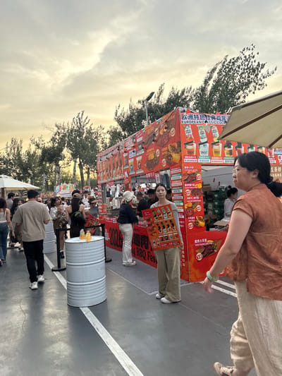 In the Chaoyang District of China, an outdoor food market bustles with activity under a cloudy sky. A large red food stall dominates the center, displaying various food items like "Tender Roasted Lamb Chops" and "Beef Wrap," priced at ¥58. The stall is adorned with promotional posters and graphics. Several people are browsing the offerings. In the foreground, a woman in a burnt orange top and beige pants walks past, her right hand resting on her hip. To her left, a young woman in beige pants and a white top holds up a large menu board. Nearby, two silver cylindrical barrels serve as makeshift tables, one topped with two orange drinks. A white canopy stretches over a portion of the market to the right, providing shade. Trees line the background, adding to the outdoor atmosphere. The ground is paved and marked with white lines, suggesting it might be an open area within a park or plaza. The overall scene depicts a lively evening at a food festival.