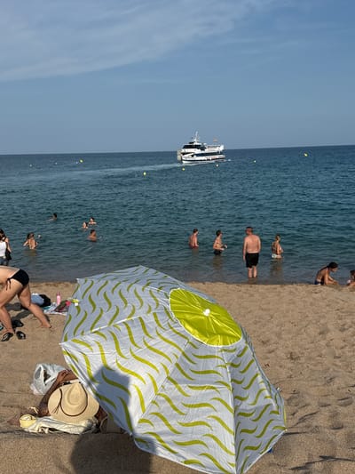 This is a photograph of a beach in Blanes, Spain, on a sunny day. The foreground of the image is dominated by a partially opened umbrella with a white and lime green wavy pattern. A person is partially visible beneath the umbrella, wearing a straw hat. A straw hat and a pink water bottle are on the sand near the umbrella. Several people are visible in the shallow water of the sea, some wading and others swimming. In the background, a white passenger boat with a blue stripe is sailing on the blue sea. The sky is a clear blue with a few wispy clouds. The water is calm and appears to be a popular spot for recreational activities.