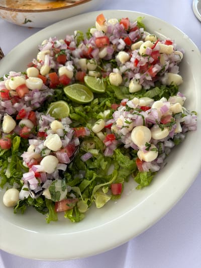 A close-up, overhead shot captures a white oval plate filled with a fresh ceviche and salad, set on a white tablecloth. The ceviche, a popular Peruvian dish, is made with large kernels of white corn, diced red tomatoes, finely chopped red onions, and chopped green cilantro. The ingredients are mixed together and mounded on top of a bed of shredded green lettuce. Three lime wedges, cut in half, are strategically placed on top of the ceviche. The lighting is bright, indicating an outdoor setting, possibly during the day. In the background, slightly out of focus, a portion of another white bowl containing a creamy, light-colored soup or stew is visible. The overall impression is of a delicious and appetizing meal, likely served in a restaurant in La Punta, Peru, a coastal district known for its seafood.