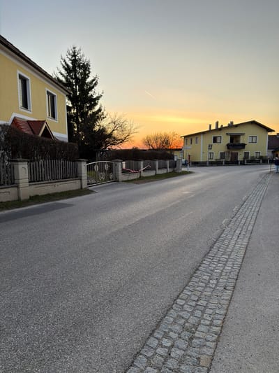 The scene depicts a residential street in Ferschnitz, Austria, during sunset or late afternoon. The sky transitions from a pale blue-grey overhead to a vibrant orange and yellow at the horizon, with a single white contrail visible in the upper-mid sky.

In the foreground, an asphalt road occupies the center, showing variations in light and shadow, indicating a low sun angle. A strip of grey cobblestones runs longitudinally along the right edge of the asphalt road.

On the left side of the road, a two-story building with a yellow facade, white-framed windows, and a brown-tiled roof is partially visible. A small, gabled porch with a matching brown roof extends from the front. In front of this building, a fence line consists of light-colored concrete posts, wooden vertical slats, and a metal pedestrian gate featuring circular decorative elements. A dense, dark hedge runs behind the fence. A tall, dark green coniferous tree stands behind the hedge. Further along the fence line, a red and white striped barrier tape is attached to a fence post and extends downward.

On the right side of the road, further into the midground, a second two-story building with a yellow facade and brown roof is visible. This building has multiple windows and a second-story balcony with dark railings. To the far right edge of the frame, a single human figure, facing away from the camera, is walking on what appears to be a paved area adjacent to the road. Bare deciduous trees are visible in the distance, silhouetted against the sunset.