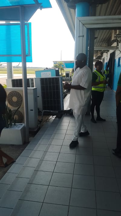 The image captures an outdoor scene at an airport in Orerokpe, Nigeria. In the foreground, a tiled walkway leads to a set of stairs. Several large air conditioning units are visible, some stacked vertically. A light blue container with the "nahco aviance" logo is also present. To the right, a man wearing a white traditional outfit and glasses stands looking to his left. Behind him, a woman in a neon yellow vest is visible. The background shows an airfield with palm trees and buildings under a bright, overcast sky. The overall impression is one of an active airport environment.