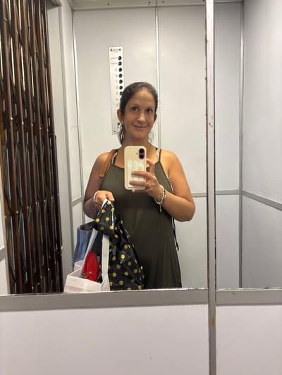 A woman is taking a selfie in an elevator. The elevator has a metal accordion gate and a control panel with floor numbers 1-9. The woman is wearing a green dress and holding a phone with a white case. She is also holding a black and gold polka-dot bag with items inside. The location is likely in Ancón, Peru, based on available data.