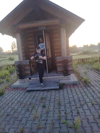 An outdoor scene in Petrovskoye, Russia, features a single-story log cabin structure with two males present. The building, constructed from horizontal logs, has a dark brown wooden roof with an overhang. A small porch area, supported by two thick wooden posts, features a concrete or brick base and a wooden step. A dark brown door is open, revealing a partially visible adult male standing inside. He wears a white t-shirt with "Reebok" text on the chest and has short, dark hair, looking towards the foreground.

A younger male, appearing to be a pre-teen or early teen, is in motion, stepping off the wooden porch step onto the paved ground. He wears a black t-shirt with "BELIEVE" and other graphics in white, dark pants, and light-colored sneakers. His arms are bent, and his hands are held in front of him, appearing to grasp indistinct dark objects, possibly gloves or a small item. He looks directly at the camera.

The ground in front of the cabin is covered with grey interlocking concrete pavers, with a reddish-brown section near the steps. Weeds and grass are visible growing in the gaps between the pavers. To the left and right of the paved area, and extending into the background, is overgrown green grass and vegetation. A wooden picket fence, dark in color, runs horizontally across the mid-ground. Beyond the fence is an open, grassy field. The background sky is bright white, suggesting diffused sunlight from a low sun, indicative of early morning or late evening.