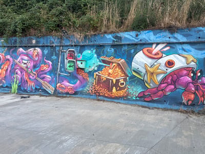 This image depicts a vibrant graffiti mural on a blue concrete wall, situated in the coastal town of Lozenets, Bulgaria. The artwork features a fantastical underwater scene with cartoonish characters. On the left, a large, smiling purple octopus with multiple tentacles holds a wooden oar and appears to be rowing. To its right, a traffic light with green and red signals, each containing a silhouette of a fish, is mounted on a pole. In the center, an open treasure chest overflows with gold coins. To the right, a large, pinkish-purple crab with its claws raised is depicted wearing a helmet adorned with a yellow starfish and orange accents. The background of the mural is painted in shades of blue, suggesting water, with stylized fish silhouettes and bubbles. Above the wall, dense green foliage and dry grasses are visible, indicating the natural environment surrounding the urban art. The ground in front of the mural is a gray concrete surface.