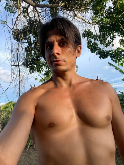 A shirtless man is shown from the chest up, standing outdoors. He has dark hair, fair skin, and a muscular build. He is looking towards the camera with a surprised expression. Two sharp, conical objects are positioned on his left shoulder. The background is dominated by lush green foliage and a blue sky with scattered clouds. The location is identified as Los Velásquez, Venezuela.