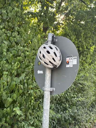 The media file shows a close-up of a bicycle helmet attached to a metal pole in front of a dense green foliage. The helmet is white with black vents and appears to be secured to the pole with a strap. A gray circular sign is visible behind the helmet, also attached to the pole. This sign has several stickers on it. One sticker, partially obscured by the helmet, reads "max jacob" and shows a CE mark with "2016" below it. Another sticker depicts a red triangle with a white line. A small sticker with a biohazard symbol is also visible. The foliage is lush and appears to be tree leaves or dense shrubbery. Sunlight filters through the leaves from the left side of the frame, suggesting it is daytime. The overall scene is outdoors, with the greenery indicating a natural or park-like setting. The context provided indicates the location is Munich, Germany. No people or specific events are depicted, but the helmet suggests cycling activity.