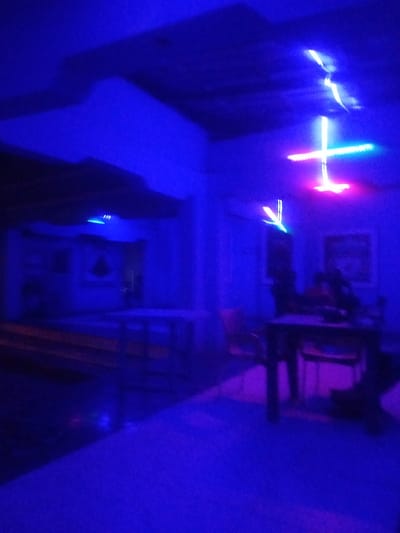 The image depicts a dimly lit interior space, likely a bar or restaurant, bathed in intense blue and purple neon lighting. The scene is blurry, suggesting motion or low light conditions. In the foreground, tables and chairs are visible, some occupied by indistinct figures. The background shows walls with framed pictures, and overhead, decorative neon lights in a cross shape emit bright white and colored light. The overall ambiance suggests a nightlife setting. The provided location of Sabon Gari Nassarawa, Nigeria, places this scene within a specific urban context.