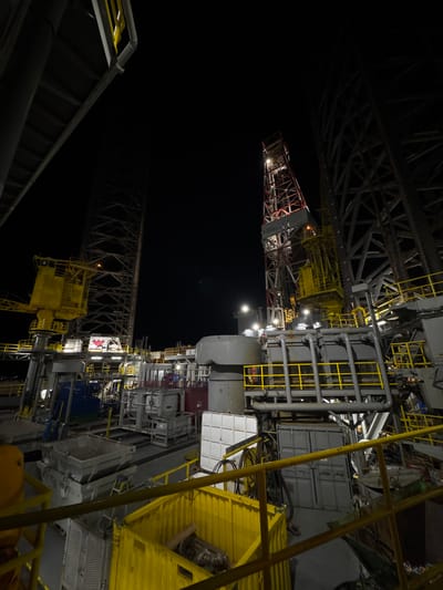 The image depicts a nighttime view of an industrial facility, likely an offshore drilling platform or a large shipyard. Towering structures, illuminated by spotlights, dominate the scene. A prominent red and white drill rig, lit from within, stands tall in the background. Surrounding it are various metal platforms, railings painted in bright yellow, and industrial containers, some white and some gray. Several lights are visible throughout the platform, casting a glow on the metal surfaces and illuminating pathways. The overall impression is one of an active, complex industrial operation at night. The location is identified as Unknown, Italy, but no specific city is mentioned. There are no people visible in the image.
