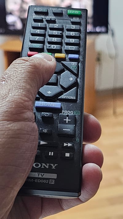 A close-up, eye-level, full shot captures a hand holding a black Sony TV remote control. The remote's buttons are visible, including number keys, directional arrows, and function buttons like "HOME" and "OPTIONS." The thumb of the hand is pressing a yellow button. The background is blurred but suggests an indoor environment with some light-colored wooden furniture and a television screen. The image does not provide any information about the city of Aksay, Kazakhstan.