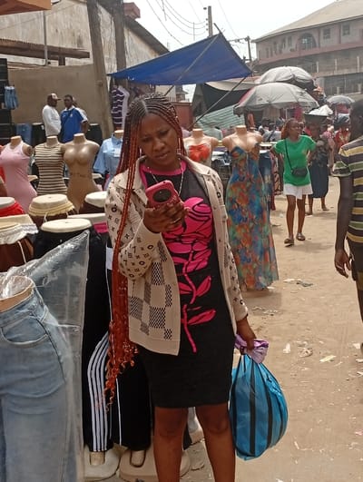 The image displays an active outdoor market scene in Asaba, Nigeria. A woman with long reddish-brown braided hair stands centrally in the foreground, looking intently at a pink smartphone held in her right hand. She is dressed in a black, form-fitting dress adorned with a prominent pink floral and leaf design, topped with a beige and white checkered cardigan. In her left hand, she carries a blue striped plastic bag containing a purple fabric item.

Surrounding the woman are numerous mannequins displaying various garments, including a pink sleeveless dress, a striped top, a nude torso form, a light blue collared shirt, a multi-colored floral strapless dress, light blue jeans, and black track pants. Several drums are visible on display stands in the lower-left.

In the mid-ground and background, multiple individuals are present. To the left, two men are standing near clothing displays. To the right, a woman in a green t-shirt and white shorts, carrying a black crossbody bag, is visible, along with other market-goers. A man in a striped shirt is partially visible on the far right.

Overhead, blue tarpaulins and large market umbrellas provide shade. The ground is a dirt or sandy surface, showing some scattered refuse. Older, multi-story buildings, characteristic of urban commercial areas, are visible in the background, with exposed electrical wiring present. The scene conveys a bustling commercial environment.