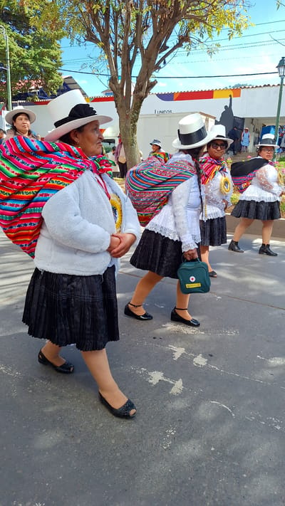 Four women wearing traditional Andean attire, including wide-brimmed white hats and colorful woven shawls, walk down a paved street in Wanchaq, Peru. The woman in the foreground on the left has a white fuzzy jacket and a black pleated skirt. The woman next to her in the center is wearing a white lace blouse and a black pleated skirt, carrying a small green medical bag. To her right, another woman is dressed similarly, and the fourth woman on the far right wears a black hat with white trim. They are walking in the same direction, suggesting a procession or parade. The background features buildings with a mural depicting a figure with an outstretched arm and the word "UNIDOS". A tree with green foliage and a bright blue sky with scattered clouds complete the scene.