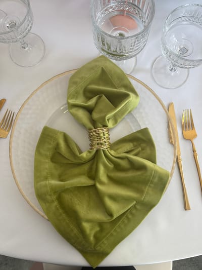 The image depicts a formally set table. At the top left are three stemmed glasses. Towards the top center is a gold-colored fork and knife. In the center is a clear textured glass charger plate. On top of the plate sits a green velvet napkin folded into a bow shape and secured by a gold metal napkin ring. At the bottom left is a third stemmed glass and at the bottom edge a gold-colored fork is visible. The table has a white linen tablecloth. The background is out of focus. There is no indication of this image being in Юг, Russia.