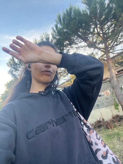 A selfie taken outdoors on a sunny day shows a person with long, dark hair covering their left eye with their right hand. They are wearing a dark grey hooded sweatshirt with the word "carhartt" embroidered in black on the front. A leopard print scarf or fabric is visible peeking out from under the sweatshirt on the right side. The person is also wearing earbuds. The background features a tall pine tree, a stone building with a red tile roof, and dry vegetation on the ground. The sky above is blue with some light clouds. The scene is consistent with an outdoor setting in Sinemorets, Bulgaria, known for its natural beauty and coastal landscapes.