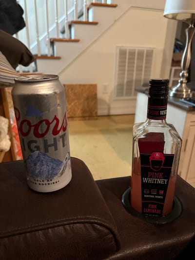 A can of Coors Light beer rests on a brown leather armrest next to a bottle of "Pink Whitney" pink lemonade vodka. The can displays the iconic Coors Light logo and the phrase "Cold As The Rockies." The vodka bottle is mostly full, with the label clearly displaying "Pink Whitney" and "Barstool Sports." In the background, a staircase, a lamp, and a kitchen counter with appliances are visible.