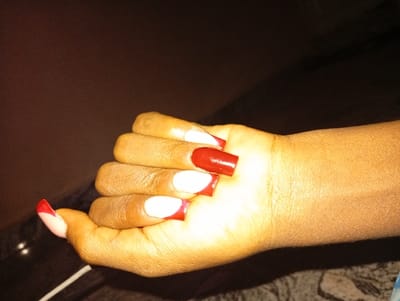 A close-up photograph depicts a human right hand, exhibiting a light brown skin tone, positioned with fingers slightly curved. The hand features long, artificial fingernails. The thumb nail displays a French tip style with a light pink base and a red tip. The index, ring, and pinky finger nails each have a white base with a red tip. The middle finger nail is painted entirely in solid red. The immediate background is dark brown and indistinct. A small section of patterned fabric, in shades of grey and white, is partially visible in the lower right quadrant. The hand is brightly illuminated, contrasting with the darker background. No visual information within the image identifies a specific location in Dabawa, Nigeria.