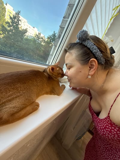 The image shows a woman and a cat at a windowsill in an apartment in Stary Oskol, Russia. The woman, wearing a red floral dress and a grey fuzzy headband, is leaning towards the ginger cat, touching noses. The cat is lying on the white windowsill, looking out a window that reveals a city view with apartment buildings and trees. The room is lit by natural light coming from the window.