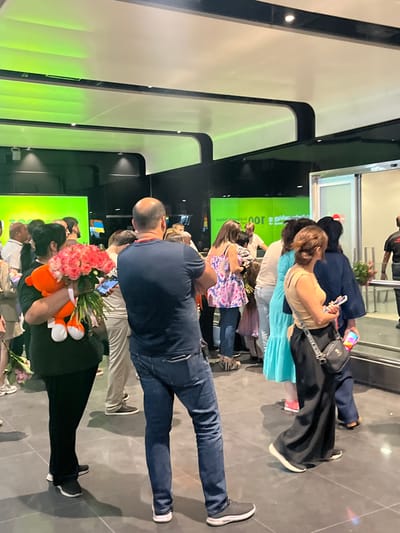 The image shows a group of people standing together, some are taking photos with their phones. One person is holding a bouquet of pink roses and an orange stuffed animal. The scene appears to be indoors, with a modern aesthetic featuring black and white accents and green lighting. There is a large green screen in the background with text that reads "100" in large white letters and some unreadable text below it in a darker color. The overall atmosphere seems to be one of celebration or anticipation. The lighting suggests it might be evening or indoors with artificial lighting. The presence of people taking photos and holding flowers indicates a special occasion or a welcoming event.