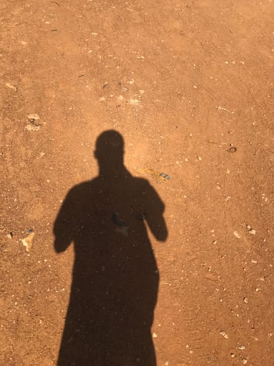 The image presents an overhead perspective of a person's shadow cast onto a sun-drenched, unpaved dirt ground in Mandate Estate, Ilorin, Nigeria.
The primary subject is a dark, elongated human shadow, positioned prominently from the lower right to the upper left of the frame. The shadow clearly outlines a head, shoulders, and arms, with one hand appearing slightly raised or extended. This distinct and lengthy shadow indicates that the photograph was taken during either the early morning or late afternoon, when the sun is low in the sky. The bright and even illumination of the ground outside the shadow suggests clear, sunny weather.
The ground itself is a light to medium brown, composed of fine soil mixed with numerous small white and grey pebbles and stones. Scattered sparsely across the surface are various small pieces of debris, including tiny fragments of what appears to be plastic or fabric (one notably blue and yellow near the center), and small organic specks.
No specific activity beyond the presence of the photographer's shadow is depicted. The scene is a simple observational shot of an outdoor setting, typical of unpaved pathways or open areas. There is no visible text in the image.