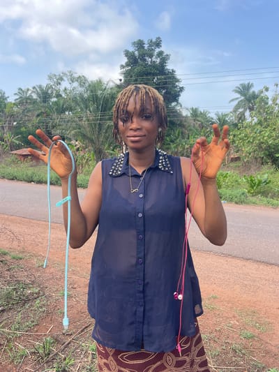 A woman stands on the side of a paved road in Ogbe, Nigeria. She wears a navy blue, semi-sheer button-down shirt with silver studs on the shoulders, and a patterned skirt. She holds a light blue USB cord in one hand and pink wired earphones in the other. The background shows a roadside with vegetation and trees, with a cloudy sky above. Power lines are visible overhead.