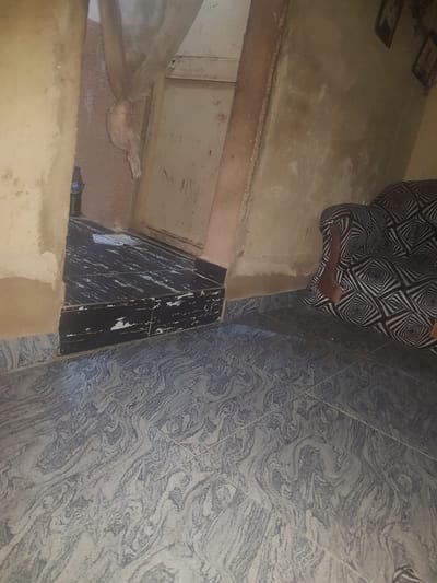The image shows a view of an interior space in Hanwa, Nigeria. A person wearing patterned black and white pants is partially visible. The floor is tiled with a grey, swirled pattern. Adjacent to the tiled floor is a section of flooring covered in black and white tiles. A wall with tan paint is visible. There appears to be a metal pipe in the bottom left corner. The overall lighting is dim.
