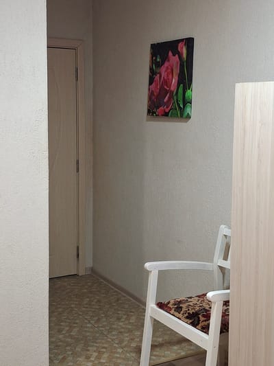 A tilted, eye-level shot captures an interior scene. A painting with pink roses is affixed to the white wall. A light-colored wooden table is partially visible at the top, with a white chair angled toward it. Part of a door with a light wood grain and a white wall below it are visible at the bottom. The background includes a tiled wall in shades of beige and brown. The location is an indoor setting in Krumovgrad, Bulgaria.