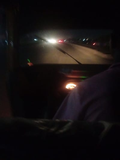 The image shows a blurry, night-time view from inside a vehicle, likely a car or a bus, as it travels on a road. The road ahead is visible, with headlights and taillights of other vehicles creating streaks of light. The interior of the vehicle is dark, with some faint lights from the dashboard visible. A person's shoulder and head are visible on the right side of the frame, suggesting they are the driver or a passenger. The overall scene is dark and somewhat indistinct due to the low light conditions and motion blur. There are no discernible landmarks or environmental details that would pinpoint the location to Sabon Gari Nassarawa, Nigeria.